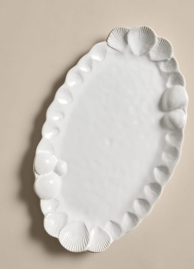 A white, oval ceramic platter with a scalloped edge featuring decorative seashell shapes, perfect for serving fresh favourites during salad season, placed on a beige background.