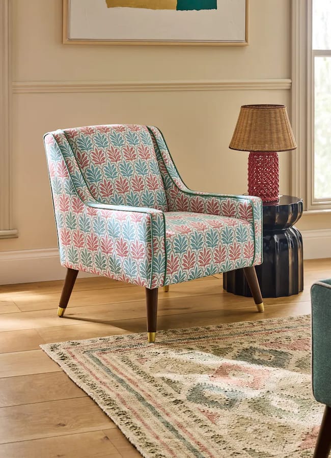 Colourful patterned armchair with wooden legs beside a red lamp on a small black table, in a cosy, sunlit room with a rug and framed artwork on the wall, echoing the vibrant aesthetic often found in maximalist hotels.