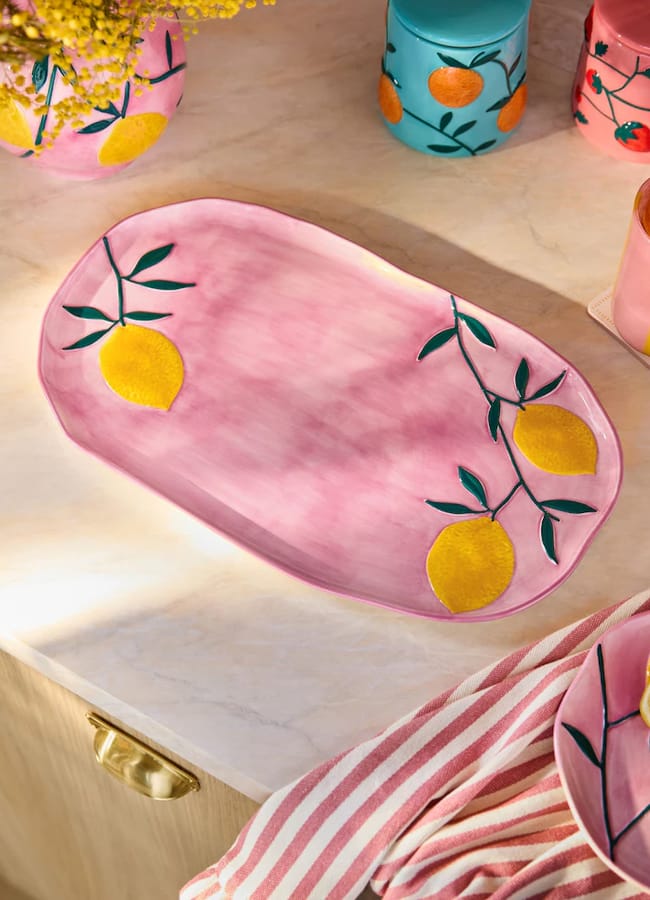 A pink ceramic platter with yellow lemons and green leaves painted on it sits on a marble worktop near a striped pink towel and colourful jars with fruit designs, making it a charming addition to any collection of kitchen accessories or foodie homewares.