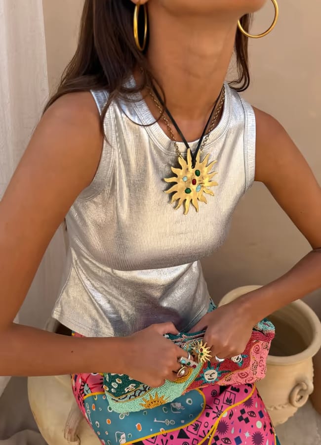 A person wearing a shiny silver sleeveless top, colourful skirt, gold hoop earrings, and sun-shaped jewellery holds a vibrant patterned clutch bag while sitting near a pottery vessel, showcasing their love for statement accessories and colourful jewellery.