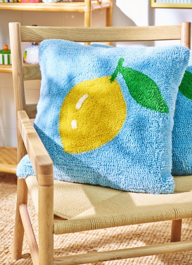 A light wooden chair with a woven seat holds a blue, fluffy cushion featuring a large yellow lemon and green leaf design—a perfect pop of colour for any foodie’s collection of homewares. Sunlight brightens the cosy, homely space.