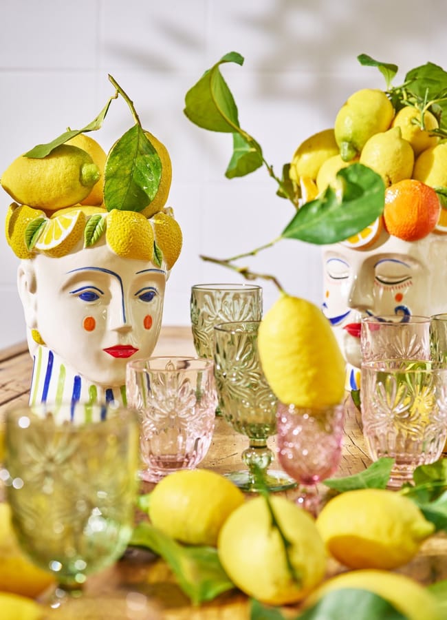 Two decorative ceramic head vases filled with lemons and green leaves sit on a table surrounded by colourful glassware, scattered lemons, and stylish kitchen accessories. The background is a white tiled wall, making this vibrant scene perfect for anyone who loves foodie homewares.