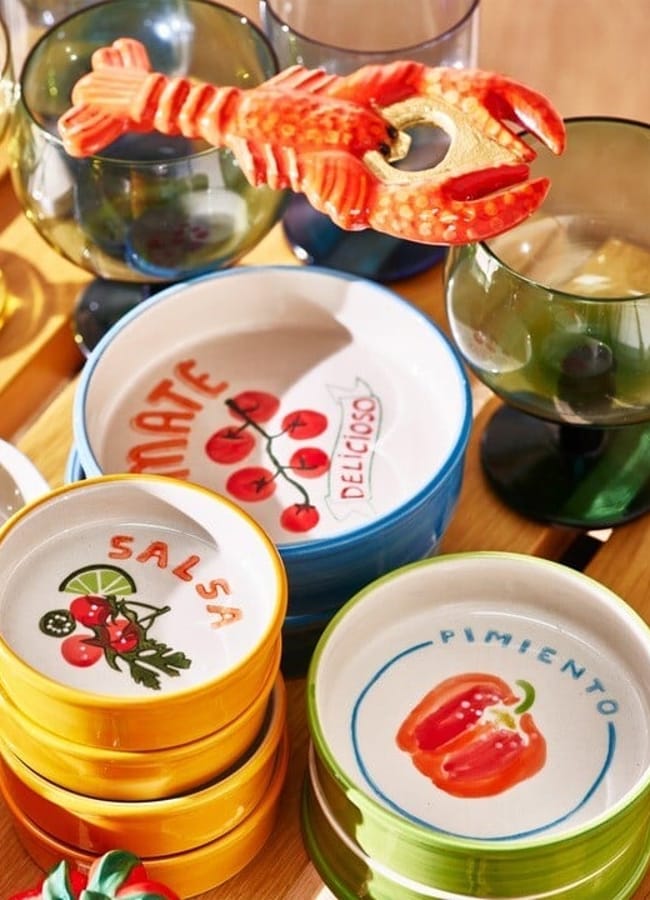 Colourful ceramic bowls with painted fruit and vegetable designs are stacked on a table, surrounded by green-stemmed glasses and a lobster-shaped bottle opener. Perfect for lovers of unique foodie homewares, the scene is bathed in sunlight that casts bright, warm shadows.