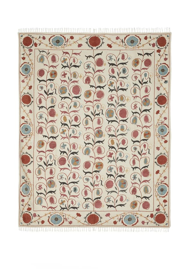 A rectangular rug with a cream background, featuring a symmetrical floral pattern in red, blue, green, and beige, bordered by a row of flowers and finishing with white tassels on both ends—perfect for adding the vibrant charm often found in maximalist hotels.