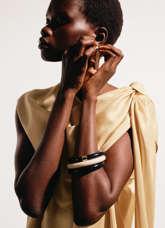A person wearing a light gold, draped, sleeveless top—an effortlessly chic nod to high street fashion—adjusts an earring. They have short curly hair and wear two chunky bracelets, one black and one white, on their wrist, against a plain background.