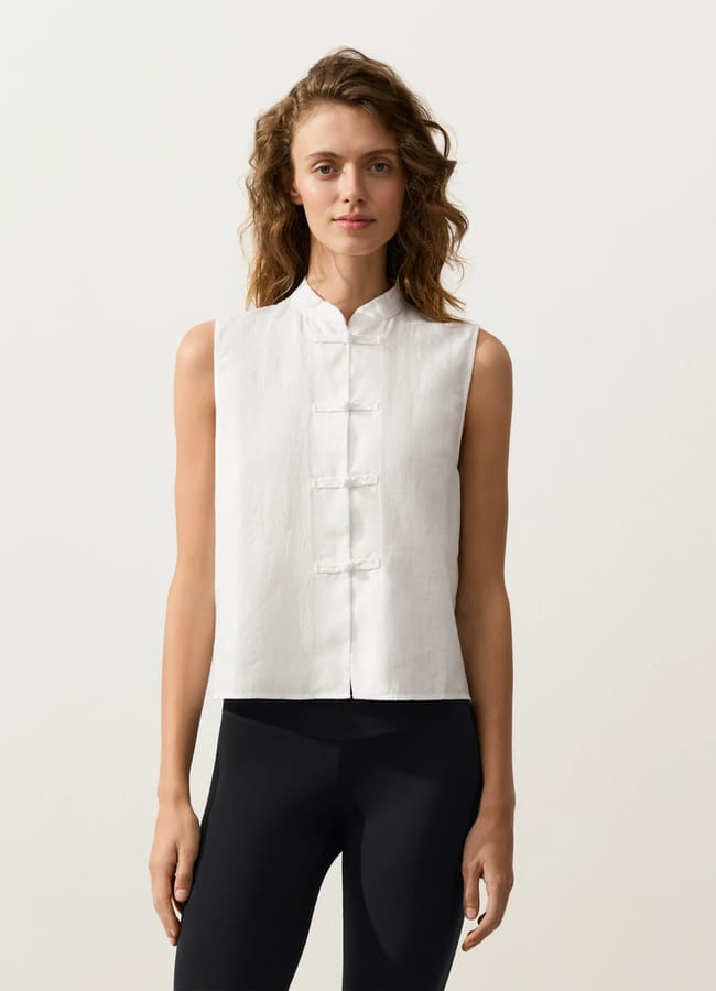 A woman wearing a sleeveless white mandarin-collar top with frog fastenings and black leggings stands facing forwards against a plain light background, showcasing subtle fashion detail for effortless outfit styling.
