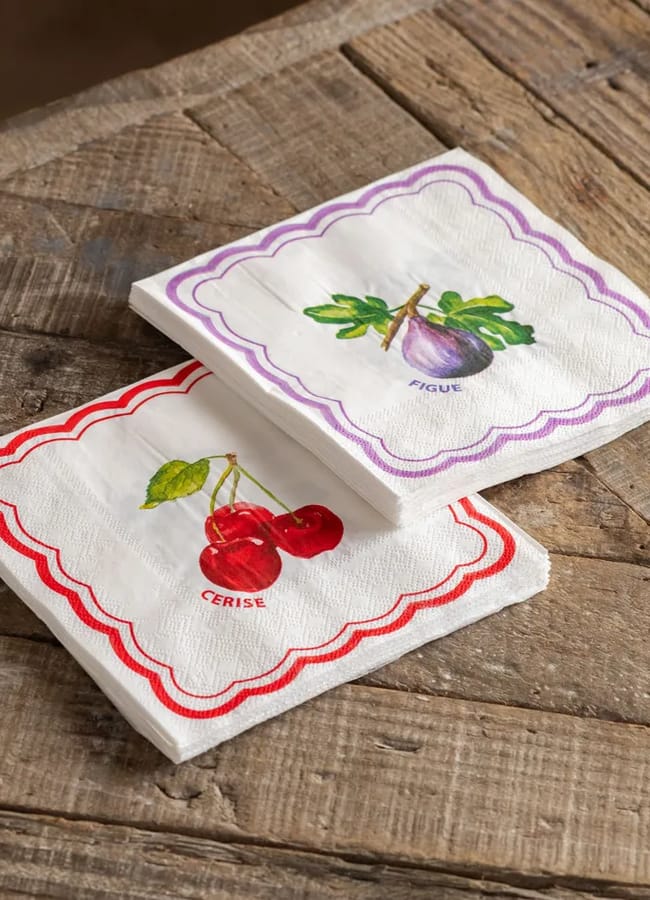Two white serviettes with scalloped borders, one red featuring cherries and labelled CERISE, and one purple featuring a fig and labelled FIGUE, placed on a rustic wooden surface—perfect homewares for the discerning foodie's table.