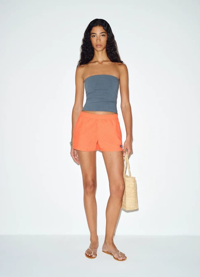A woman stands against a plain background wearing a grey strapless top, bright orange alternatives to denim shorts, clear sandals, and carrying a woven tote bag. Her hair is long and curly—a fresh take on summer fashion.