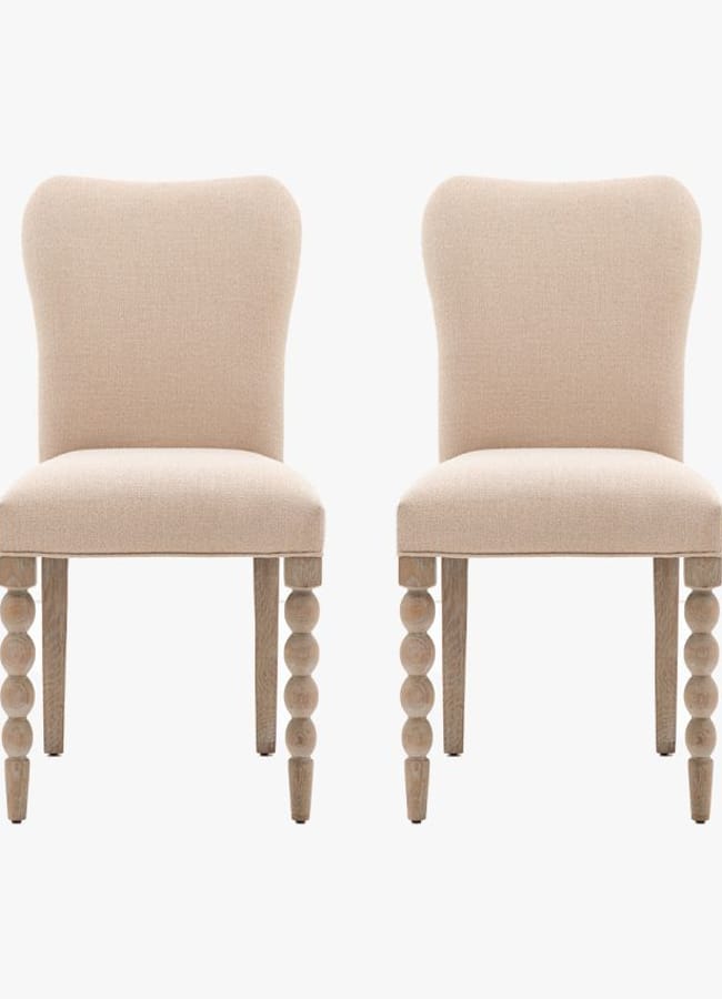 Two beige upholstered dining chairs with curved backs and unique legs featuring three round, stacked wooden spheres on each front leg evoke the playful sophistication often found in maximalist hotels, set against a plain white background.