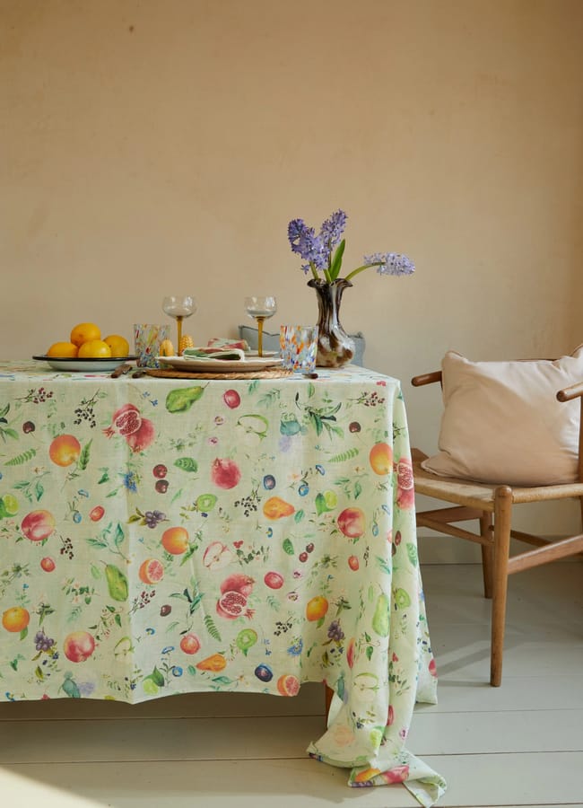 A table with a colourful fruit-patterned tablecloth, lemons, glasses, and a vase of purple flowers creates a cheerful scene. Thoughtfully arranged foodie homewares and kitchen accessories add charm, while a wooden chair with a beige cushion sits invitingly next to the table.