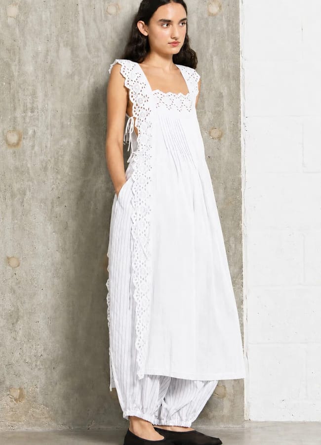 A woman stands against a concrete wall wearing a white dress with broderie anglaise details over loose, striped trousers—a nod to summer micro trends. She has long dark hair, her hands in her pockets, and is wearing black flat shoes.
