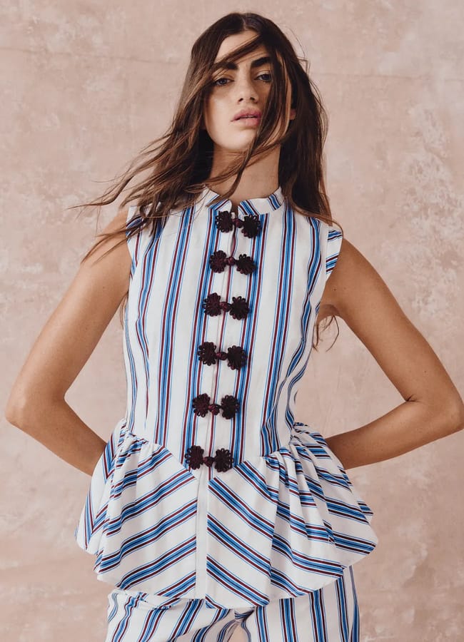 A woman poses in a sleeveless, striped peplum top with vertical blue, white, and red lines and black floral buttons, showcasing expert outfit styling. Her matching striped trousers complete the chic ensemble as she stands against a textured beige background.