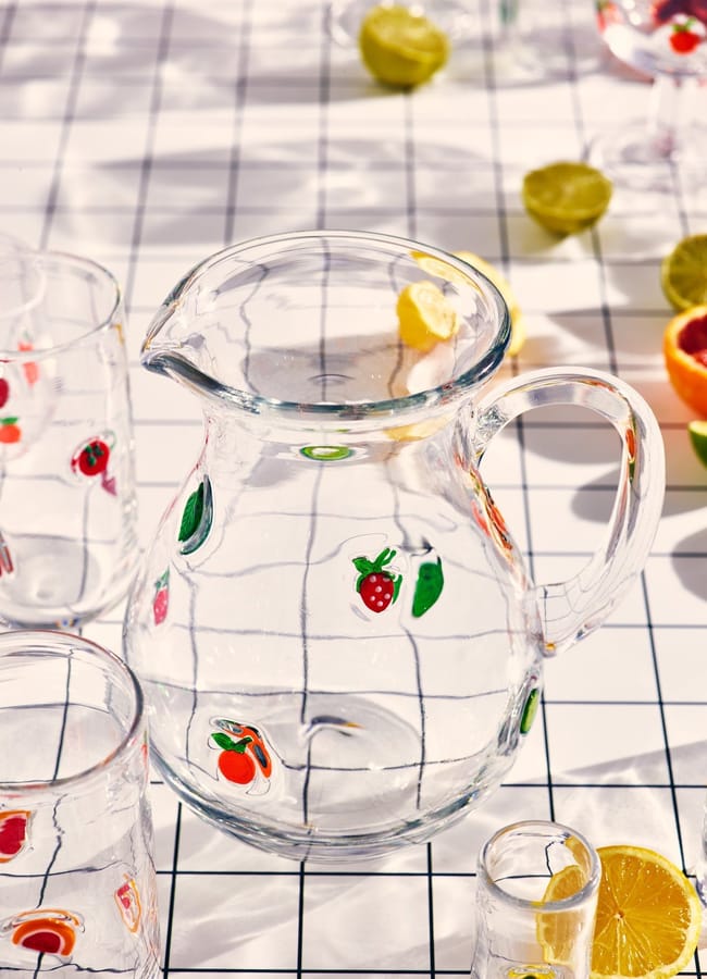 A glass jug decorated with fruit illustrations sits on a white grid-patterned table, surrounded by matching glasses and halved citrus fruits—a perfect scene for any foodie’s collection of charming homewares.