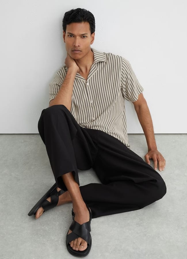 A person sits on the floor against a white wall, wearing a beige short-sleeved striped shirt, black trousers, and sleek black men's summer shoes, with one leg bent and an arm resting on the floor.