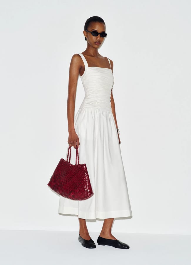 Woman wearing a white sleeveless midi dress, black flats, sunglasses, and holding a large red woven handbag, standing against a plain white background—a chic look inspired by Kendall Jenner style.