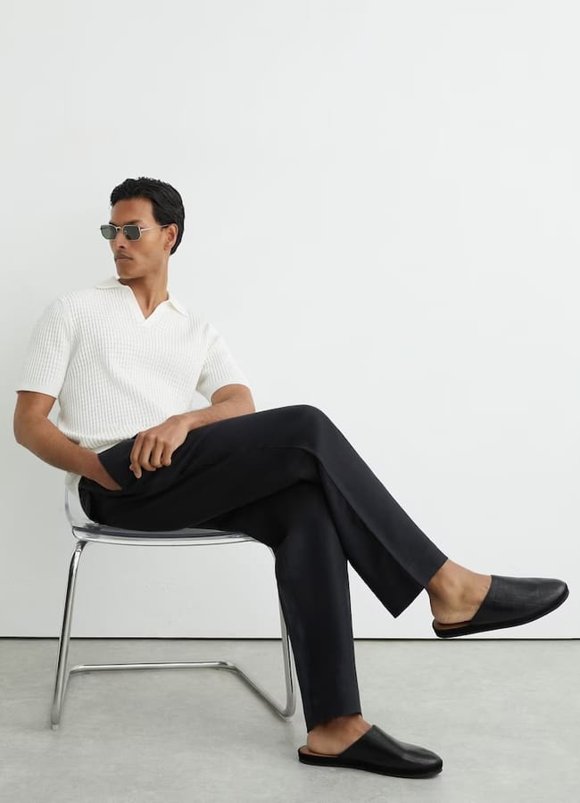 A man wearing sunglasses, a white short-sleeved knitted shirt, black trousers, and sleek men's summer shoes sits cross-legged on a clear chair, looking to the side in a minimalist setting.