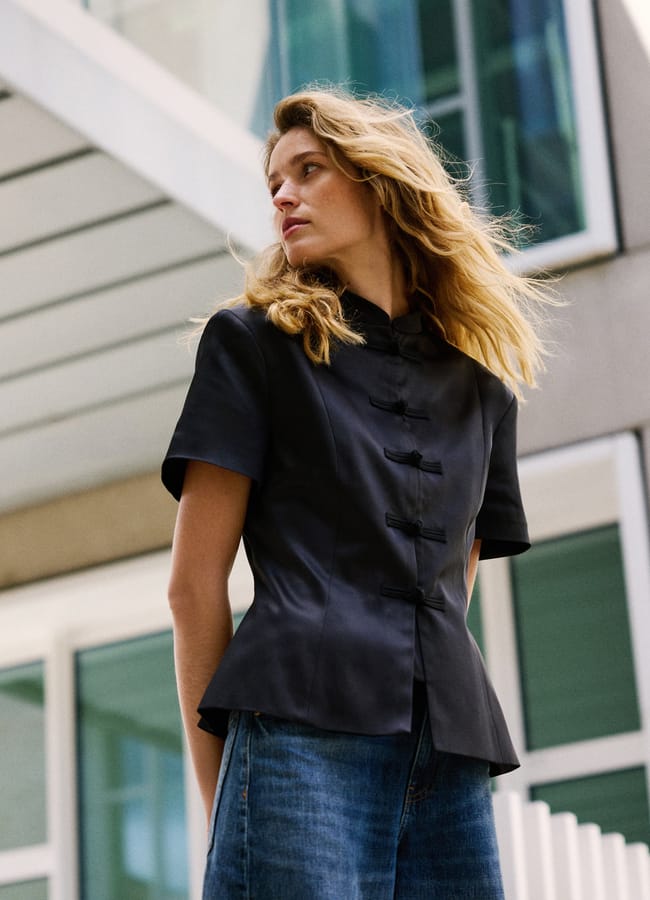 A person with long blonde hair wearing a short-sleeved black top and blue jeans stands outside in front of a modern building with large windows, looking off to the side, effortlessly embodying the latest high street fashion trends.