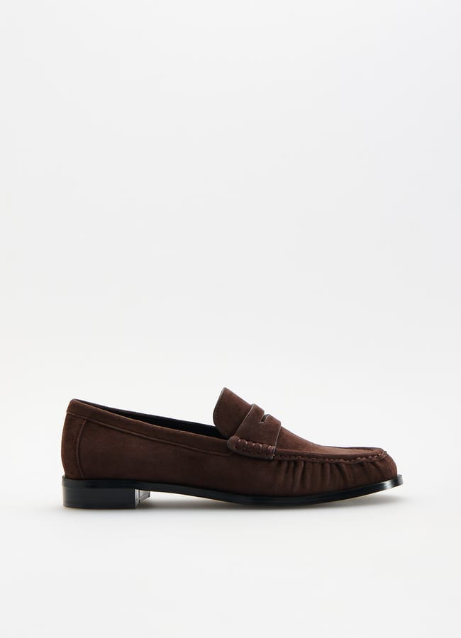 A brown suede loafer shoe with a low black heel and stitched detailing, displayed against a plain white background—effortlessly combining comfort and high street fashion style.