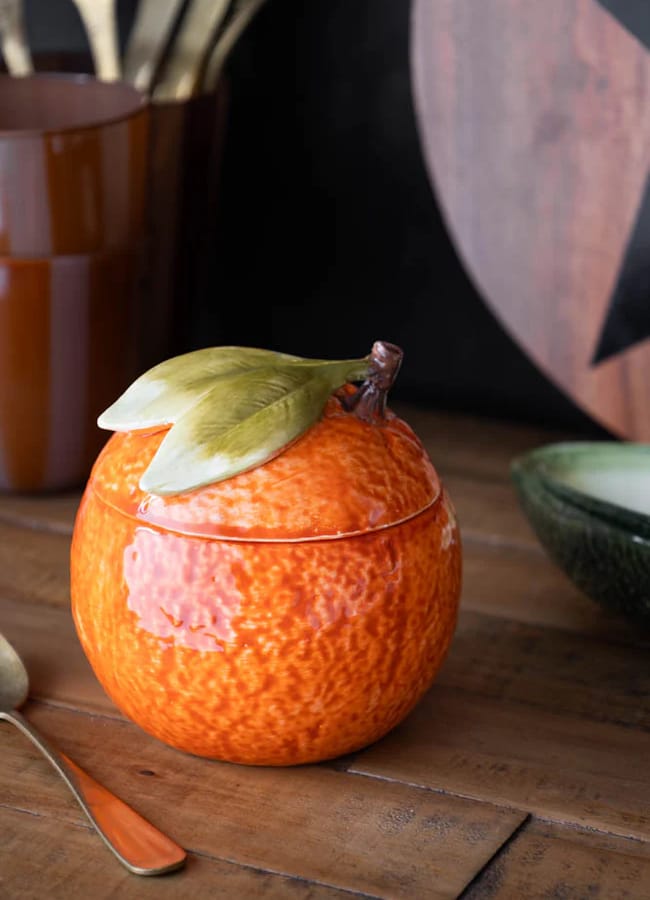 A ceramic container shaped like an orange with a lid featuring green leaves sits on a wooden table near a spoon, brown mug, and green bowl—an ideal addition to your collection of kitchen accessories.