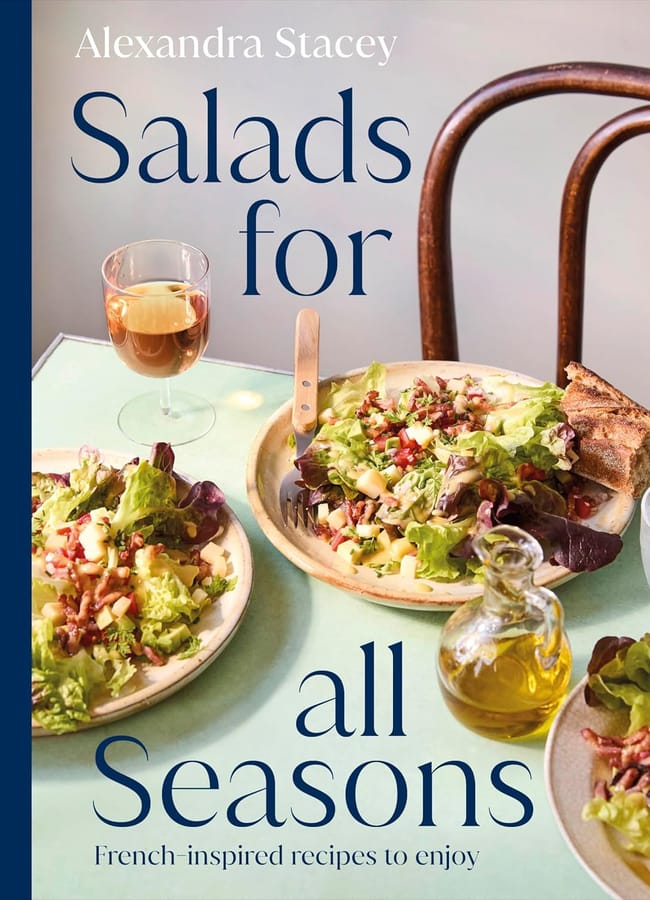 Cover of Salads for All Seasons by Alexandra Stacey, capturing the essence of salad season with plates of fresh salad featuring lettuce, apples, walnuts, bread, a glass of drink, and a small bottle of oil on a light green table.