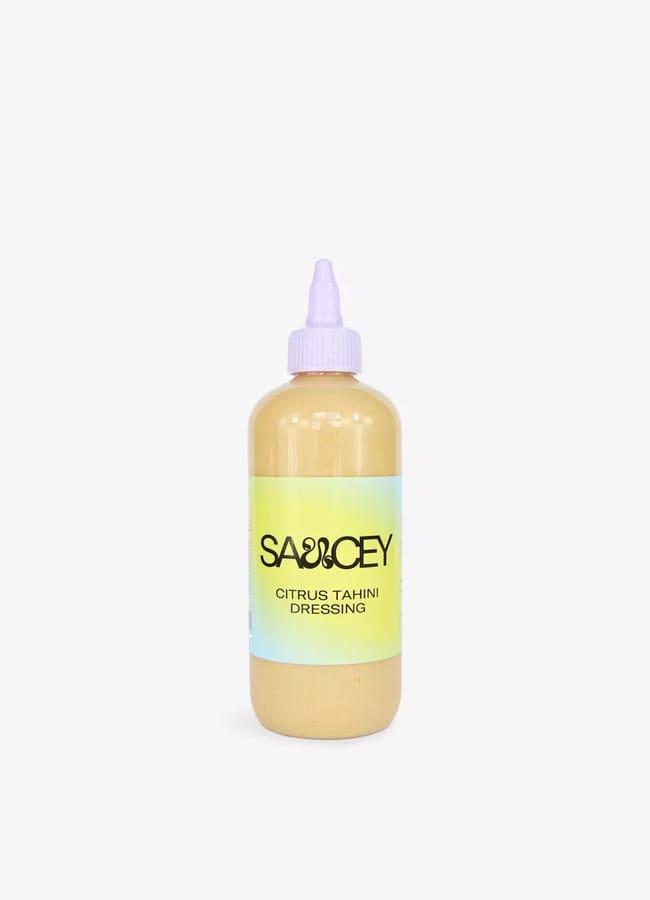 A plastic squeezy bottle with a purple cap, filled with light yellow sauce. The label reads SAUCEY Citrus Tahini Dressing on a gradient yellow-green background—perfect for brightening up salad season.