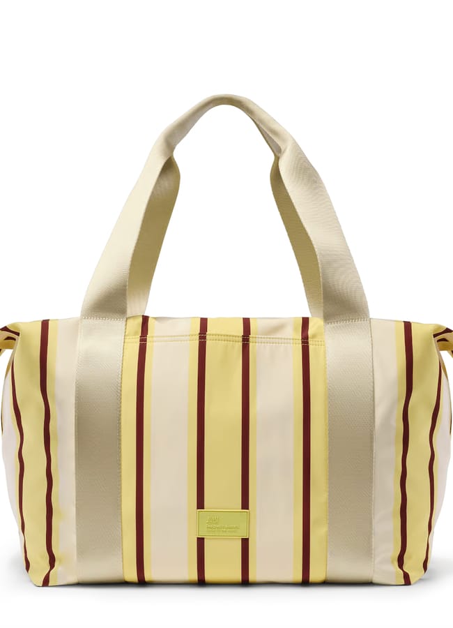 Striped holdall with yellow, cream, and brown vertical stripes, large beige handles, and a rectangular yellow label on the front. Perfect for those wondering what to wear in Ibiza, the bag has side ties and a zip fastening—ideal for carrying all your holiday essentials in style.