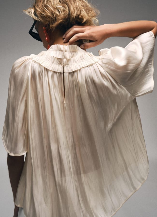 A person with short blonde hair, wearing sunglasses and a flowing cream-coloured pleated blouse—a nod to high street fashion—stands with their back to the camera, adjusting the collar.