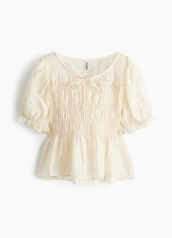 A light cream, short-sleeve blouse with a ruched bodice, puffed sleeves, a small front bow, and a frilled hem—perfect for those wondering what to wear in Ibiza—is displayed on a white background.