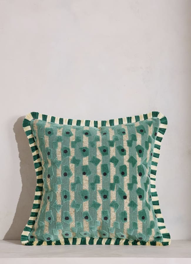 A square decorative cushion with a green and cream geometric pattern and a striped green, cream, and black border, reminiscent of the bold designs often found in maximalist hotels, placed against a plain off-white wall.