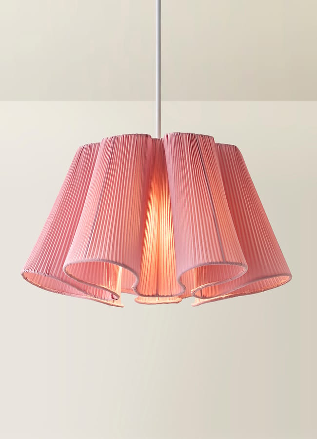A pink, pleated pendant lampshade hangs from the ceiling, softly glowing with warm light against a neutral background—an inviting touch reminiscent of the bold, curated style often found in maximalist hotels.