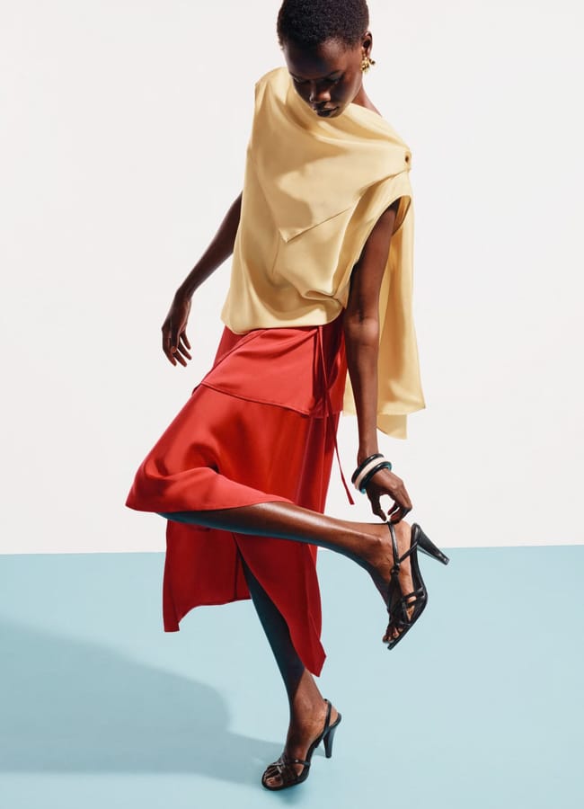 A person poses in a flowy beige top and a silky red skirt, epitomising high street fashion while wearing black heeled sandals. They stand on a blue floor with a white background, slightly leaning as they adjust a sandal strap.