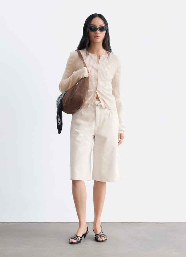 A woman wearing sunglasses, a sheer beige cardigan, cream Bermuda shorts reminiscent of the best denim shorts, black sandals, and holding a brown shoulder bag with a black bandana tied to it stands against a plain white background.