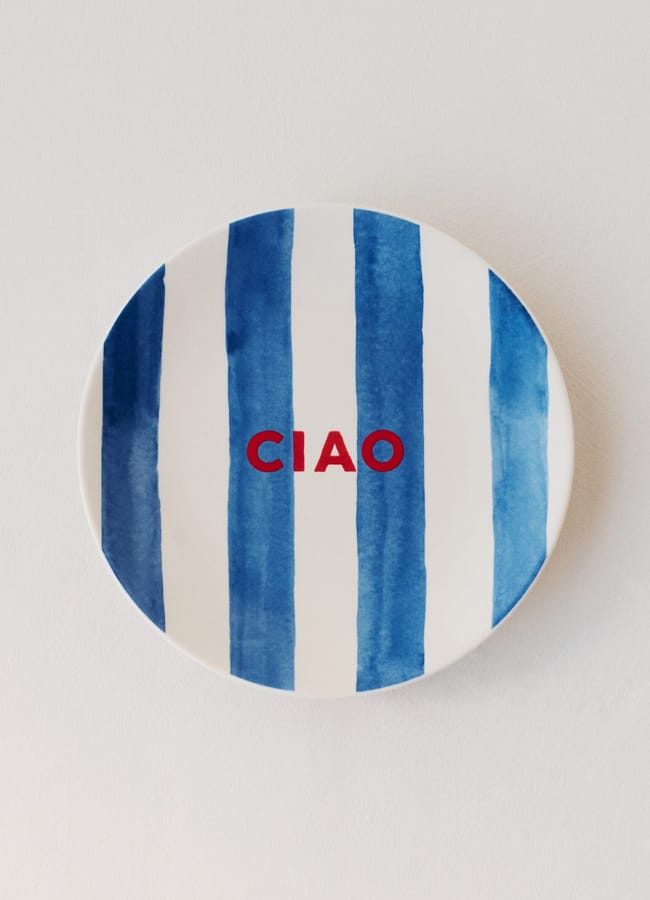 A white plate with bold vertical blue stripes and the word CIAO in red capitals at the centre, perfect for adding a fresh touch to salad season, displayed against a plain white background.