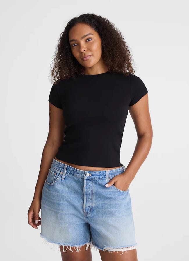 A woman with curly hair wearing a fitted black T-shirt and the best denim shorts in light blue stands against a plain white background, one hand in her pocket and a slight smile on her face.