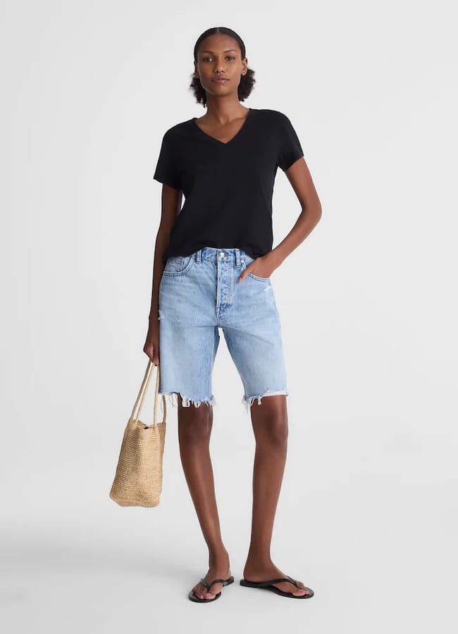 A person stands against a plain background wearing a black V-neck T-shirt, the best denim shorts in light blue with frayed hems, black flip-flops, and holding a woven straw tote bag.