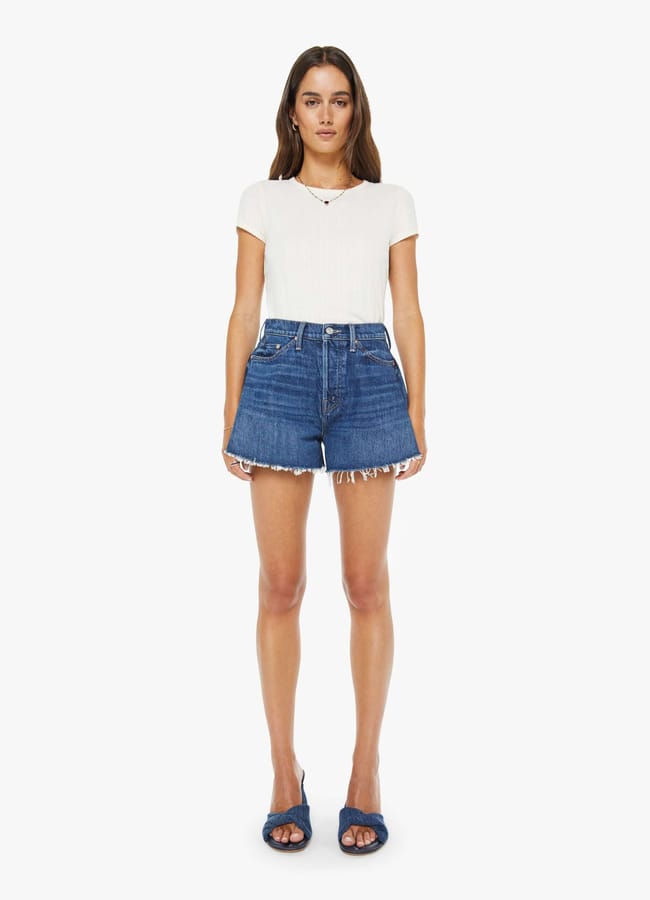 A woman stands facing forward, wearing a white short-sleeve top, blue high-waisted best denim shorts with frayed hems, and navy blue open-toe sandals. She has long brown hair and a neutral expression against a plain white background.
