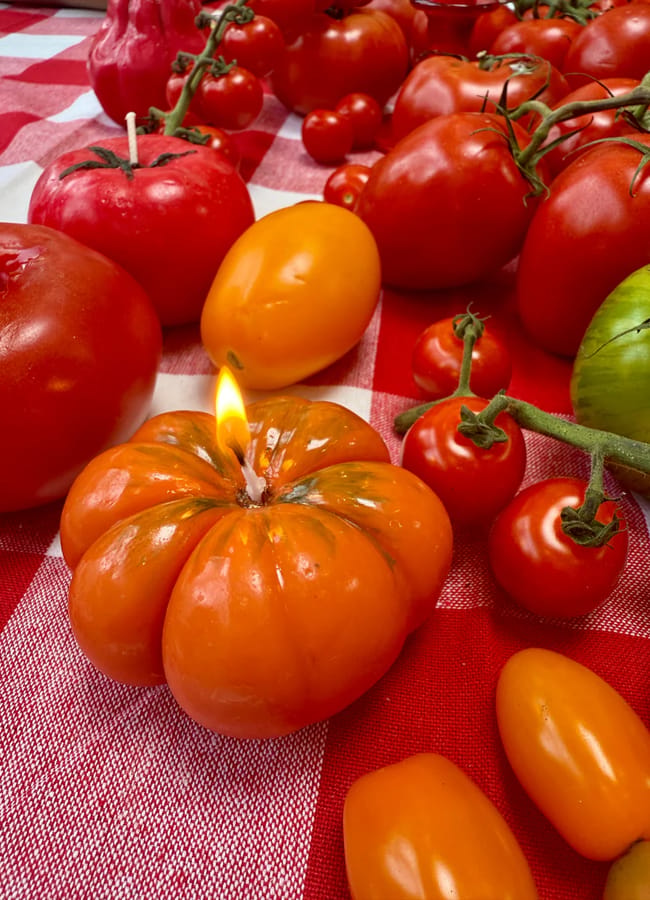 An assortment of red, orange, and green tomatoes on a red and white chequered cloth creates a vibrant foodie display; one orange tomato in the foreground is cleverly crafted as a candle, with a visible flame—an artful blend of fresh produce and whimsical homewares.