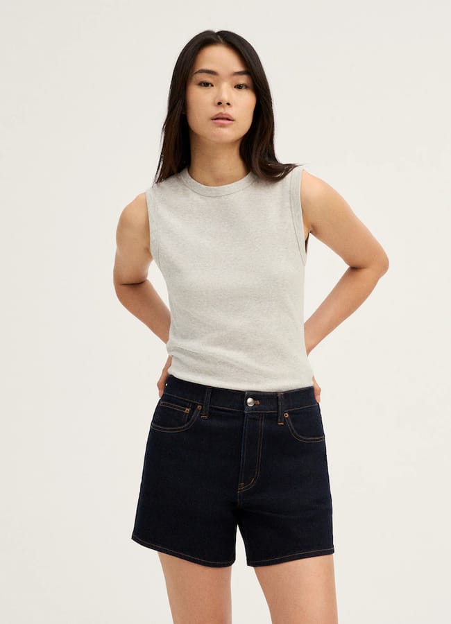A person with long dark hair wears a sleeveless light grey top and high-waisted best denim shorts, standing against a plain light background with hands resting on hips.
