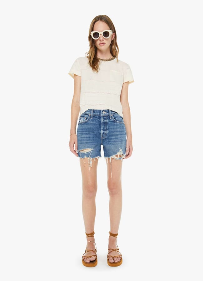A woman wearing white sunglasses, a short-sleeve cream top, best denim shorts, and brown platform sandals stands facing forward against a plain white background.