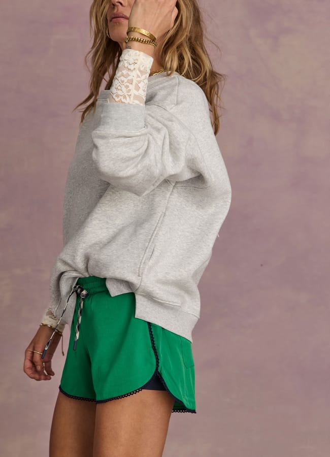 A woman in a grey sweatshirt with lace cuffs and green shorts stands against a purple textured background, showcasing one of this season’s must-see fashion trends: stylish denim alternatives.