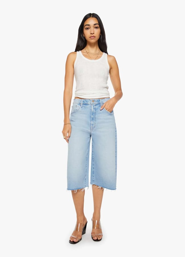 A woman stands facing forwards, wearing a white sleeveless top and the best denim shorts—light blue, knee-length with frayed hems—paired with clear high-heeled sandals against a plain white background.