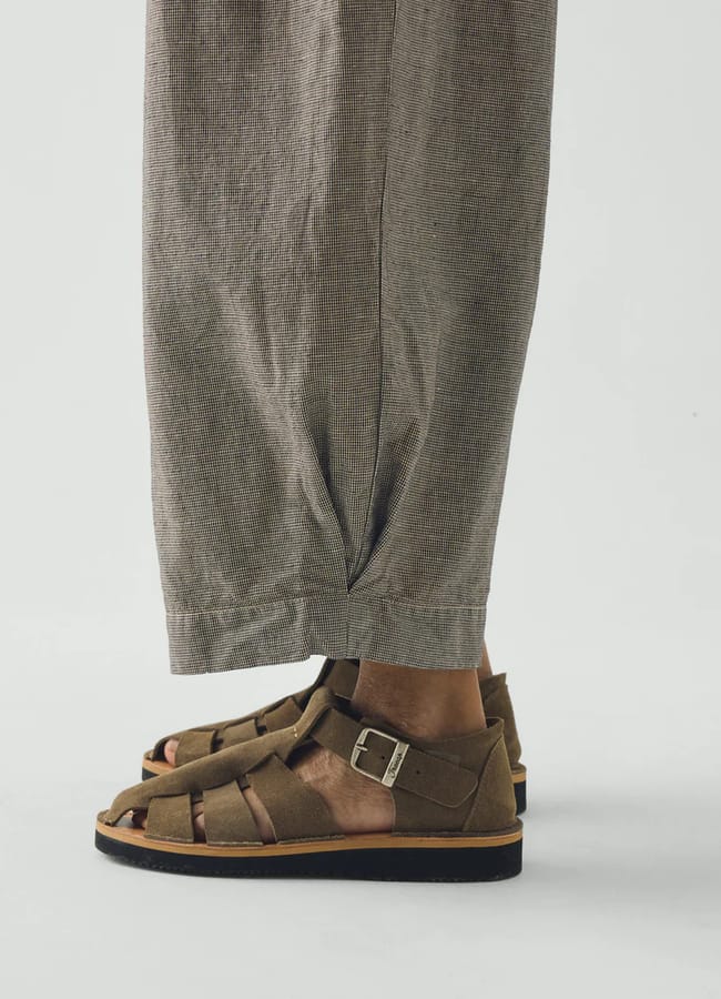 Close-up side view of a person wearing loose, textured, light brown trousers and brown leather men's summer shoes with a buckle, standing on a white background.