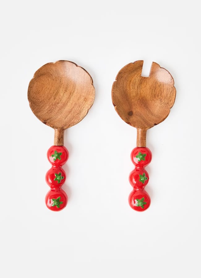 Two wooden salad servers, perfect for salad season, feature handles adorned with three red tomatoes each. One has a solid spoon, the other a forked edge, both set against a white background.