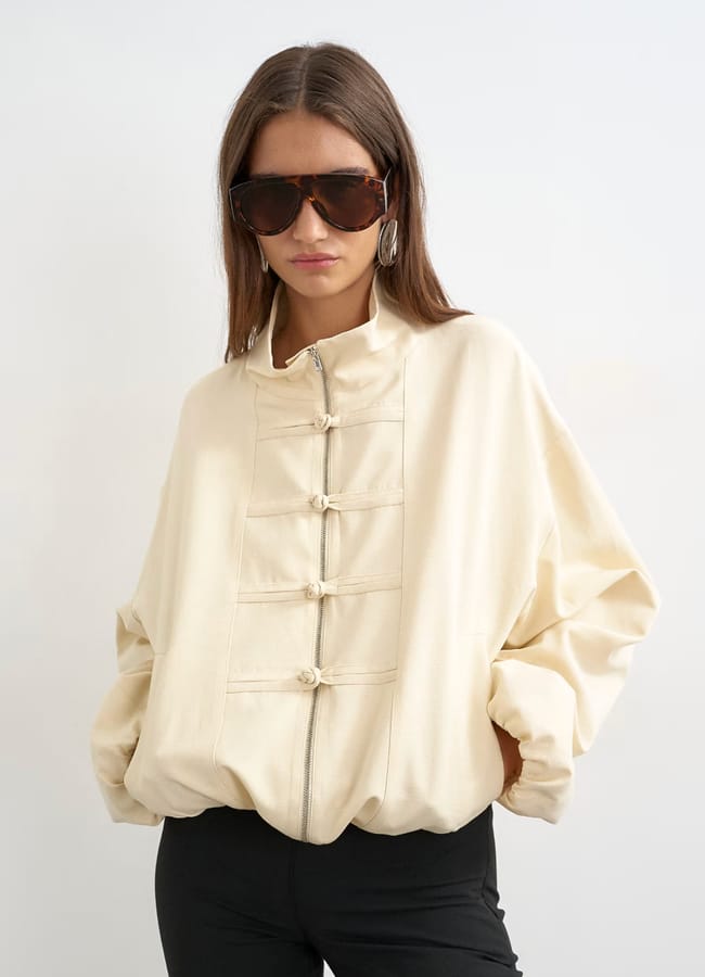 A woman wearing a cream-coloured, button-up jacket with knotted fastenings—a standout fashion detail—paired with black trousers, large dark sunglasses, and hoop earrings stands against a plain white background.
