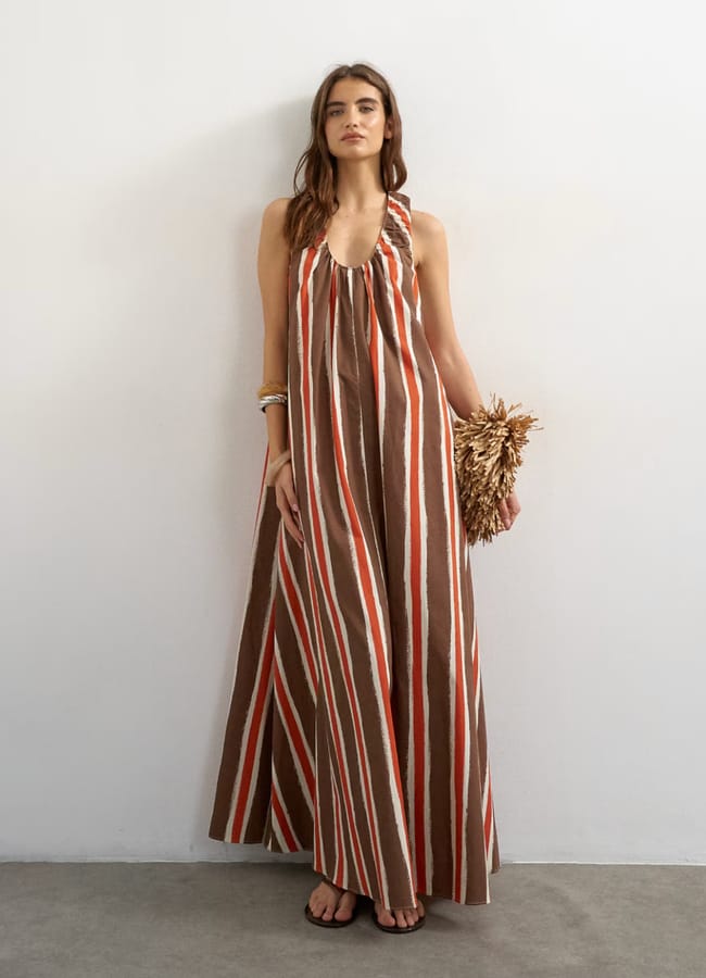 A woman in a brown, white, and red striped halter maxi dress, channelling high street fashion, stands against a plain wall whilst holding a textured clutch. She wears black sandals and bracelets, with loose hair and a relaxed expression.
