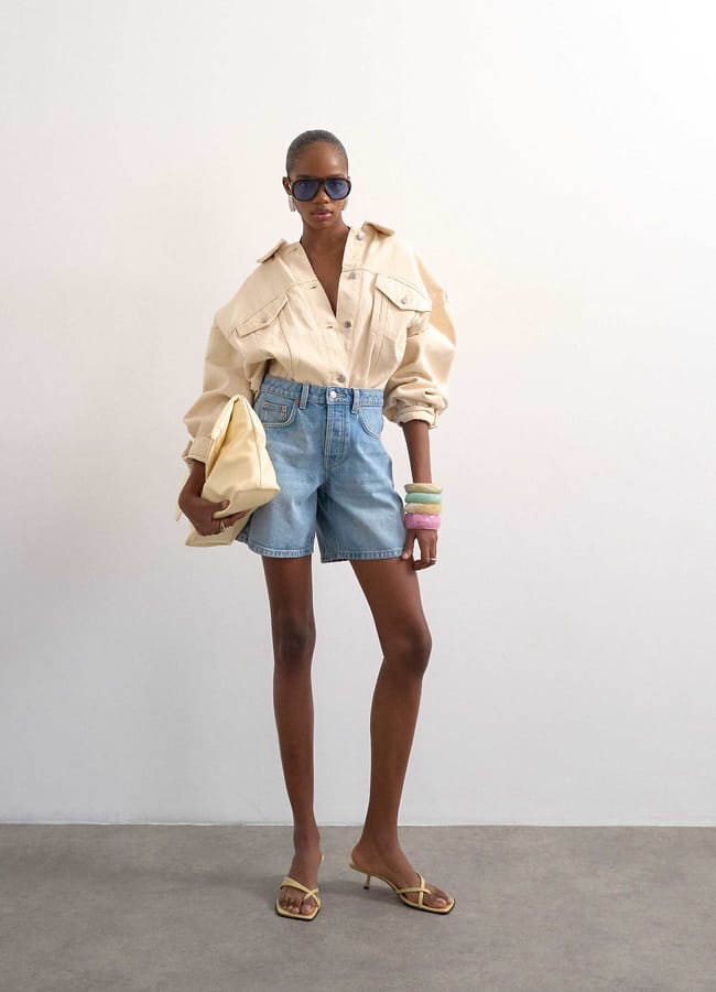 A woman wearing a beige oversized button-up shirt, best denim shorts, heeled sandals, and sunglasses stands against a plain wall. She carries a large beige clutch and wears colourful bangles on her wrist.