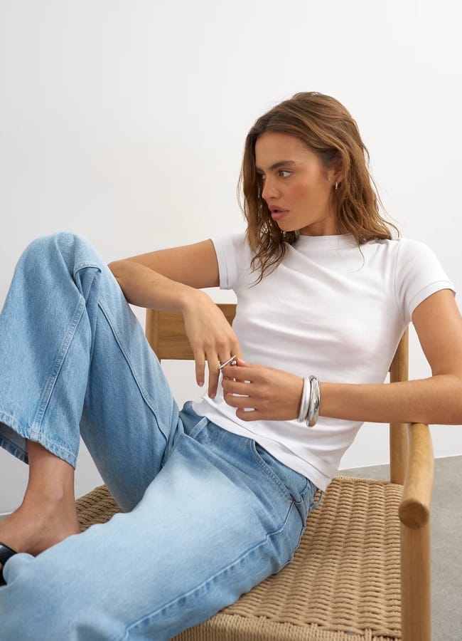 A woman in a white T-shirt and light blue jeans sits on a woven chair, looking to her left. Channelling Kendall Jenner style, she has wavy brown hair and wears large silver bangles, with one hand resting on her lap.