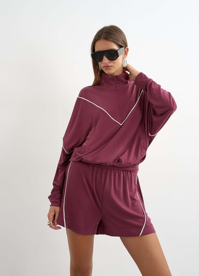A person wearing a loose maroon long-sleeve top and matching shorts with white piping stands against a plain wall, posing with one hand on their neck and wearing large black sunglasses. Perfect for those seeking effortless summer outfits or unique fashion alternatives to classic denim shorts.