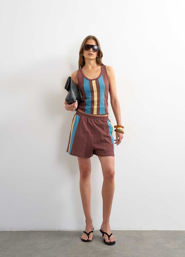 A woman in sunglasses wears a striped sleeveless top, matching alternative shorts with side stripes, black sandals, and holds a black clutch bag. She stands against a plain white wall.