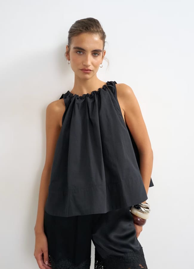 A woman stands against a white wall wearing a sleeveless black top with a gathered neckline and black lace-trimmed shorts, holding a small clutch—a chic look that captures the effortless appeal of high street fashion.
